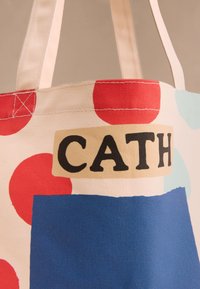 Lõuendist käekott punaste täppidega, beežide sangadega, sinise ruuduga ja julgete mustade tähtedega "CATH" beežil plaastril.