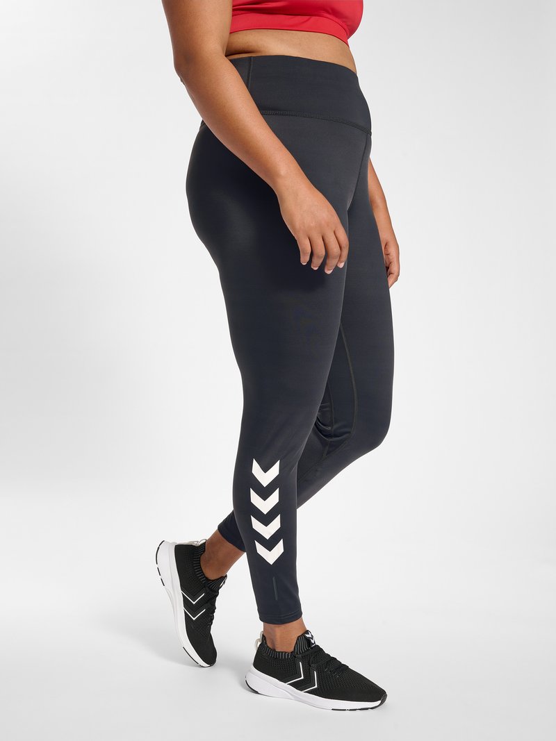 Frau trägt schwarze Leggings mit hoher Taille und weißen Chevron-Mustern an den unteren Beinen sowie schwarze Sneaker mit weißen Sohlen.