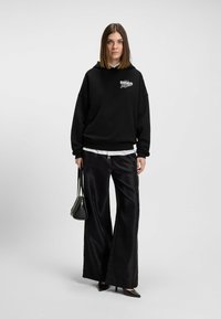 Felpa nera con stampa grafica "DISCO", abbinata a pantaloni neri in satin larghi. Il modello tiene una piccola borsa nera. Sfondo semplice.