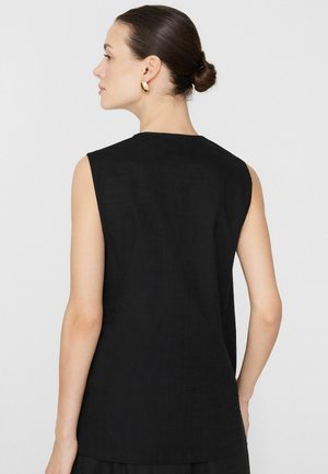 Žena s vlasy v drdolu, nosící zlaté kruhové náušnice a černý top bez rukávů, otočená vlevo, s většinou zad viditelných na bílém pozadí.