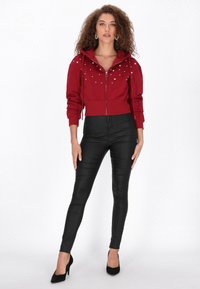 Sudadera roja recortada con cremallera y tachuelas decorativas plateadas, combinada con pantalones ajustados de cuero negro y tacones altos negros. Textura suave en general.