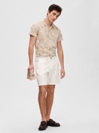 Chemise à manches courtes avec un motif de feuilles beige et blanches, associée à des shorts en lin blancs et des chaussures marron, tenant un objet beige.