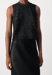 Czarny koronkowy crop top z wysokim dekoltem, misternymi wzorami kwiatowymi oraz falbaniastym dołem. Materiał ma teksturowany, przejrzysty wygląd.