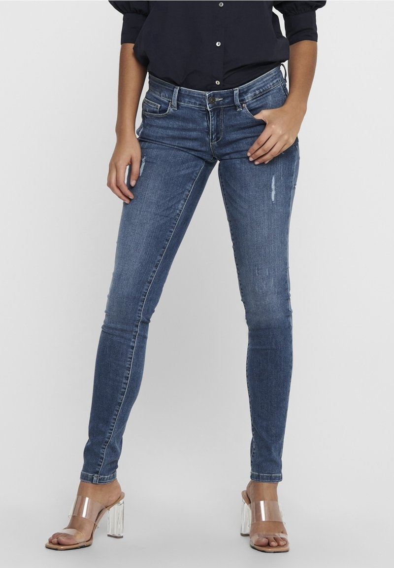 Mujer vestida con jeans ajustados azules con ligeras desgastes, una camiseta de manga larga oscura y sandalias de tacón alto transparentes, apoyada contra un fondo liso.