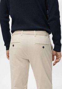 Light beige trousers with pockets featuring black buttons. The waistband is elastic. A navy knitted sweater is worn above.