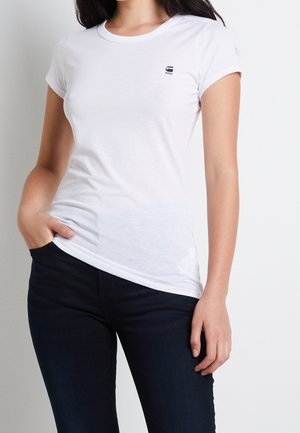 Vrouw draagt een nauwsluitend wit G-Star RAW T-shirt en donkerblauwe spijkerbroek, met haar linkerhand in haar zak tegen een effen witte achtergrond.