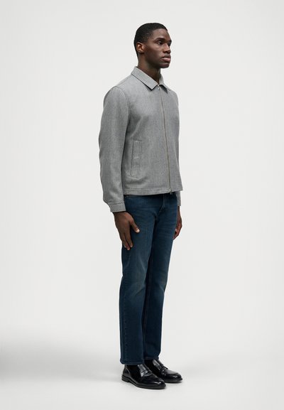 Pelēks kapuci žakets ar apkakli, divām sānu kabatām un gludu tekstūru, saskaņots ar tumši ziliem džinsiem un melnām ādas kurpēm.