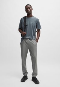 T-shirt de sport gris avec un logo subtil, pantalon gris et baskets noires. Le modèle se tient avec un sac à dos noir, sur un fond neutre.