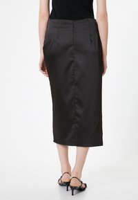 Gonna midi nera in satin con una forma aderente, chiusura con zip sul retro e pieghe sottili, indossata con sandali neri con tacco e laccetti.