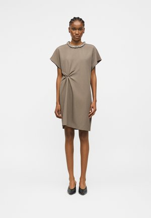 Femme debout, face à l'avant, portant une robe taupe mi-genoux avec un détail froncé à la taille et des chaussures noires à bout pointu.