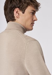 Beige Rollkragenpullover mit geripptem Kragen und strukturiertem Strickmuster, mit Schulterbahnendetails und taillierter Silhouette.