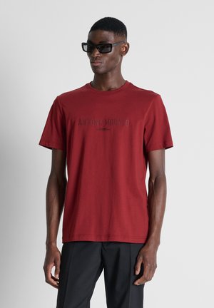Uomo che indossa occhiali da sole neri, maglietta rossa a maniche corte con testo e pantaloni neri, in piedi davanti a uno sfondo grigio chiaro uniforme.