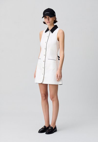 Vestido blanco sin mangas con cuello negro y detalles en negro, que presenta un cierre de botones en la parte delantera, dos bolsillos y un lazo en la parte trasera. Gorra negra y mocasines.