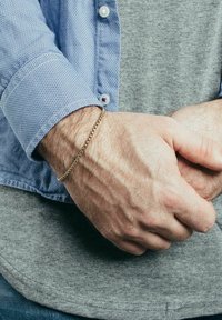 Brățară din aur pe un încheietură, texturată cu un design împletit. Încheietura este parțial acoperită de o cămașă cu model albastru.