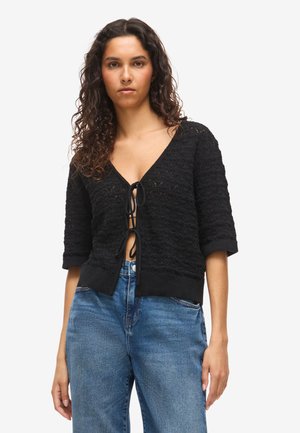 Žena s kučeravými vlasmi, ktorá má na sebe čierny pletený kardigán s viazaním vpredu a modré rifle s vysokým pásom, stojí pred jednoduchým svetlým pozadím.