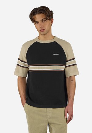 Ung mann med bølgete brunt hår iført en svart og beige kortermet genser med horisontale striper og beige bukser, stående med hendene i lommene.