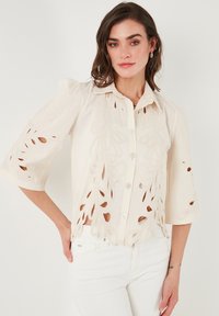 Blusa blanca con patrones de corte floral y cierre de botones en la parte delantera. Tiene mangas cortas abullonadas y una tela texturizada.