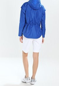 Modrá lehká bunda s vypasovaným pasem a kapucí, v kombinaci s bílými šortkami a stříbrnými teniskami. Hladká textura a neformální design.