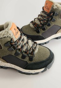 Bottes de randonnée en daim vert et mesh, doublure en fausse fourrure grise, accessoires noirs, accents orange et semelle texturée robuste.