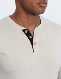 Närbild av person som bär en beige henley-tröja med svart innerspalte och tre bruna knappar, där två är knäppta och en är öppen.