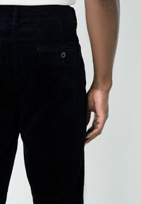 Dark navy corduroy pants with vertical ribbing. Features a back pocket with a button and a tailored shape. Smooth texture, fitted waist.
