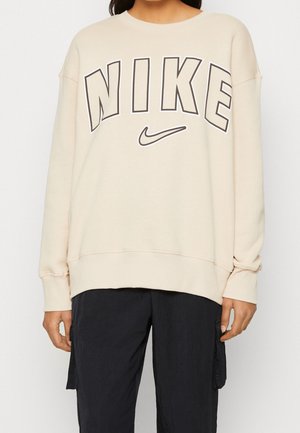 Osoba ubrana w beżową bluzę z dużym czarnym napisem "NIKE" z obrysem oraz logo Swoosh, połączoną z czarnymi spodniami typu cargo.