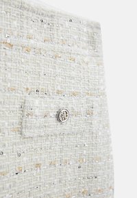 Nahaufnahme eines strukturierten Creme-Tweed-Stoffs mit Silber- und Golddrähten sowie einem kleinen runden silbernen dekorativen Knopf.