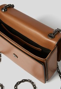 Brązowa skórzana torba na ramię o wyraźnym kształcie, z metalowymi elementami w odcieniu srebra i łańcuszkowym paskiem. Wnętrze ma czarne wykładanie oraz kieszeń na drobiazgi.