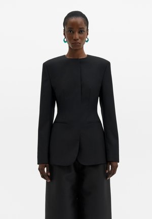 Vrouw die een gestructureerde zwarte blazer met gewatteerde schouders en bijpassende wijde broek draagt, staand tegen een effen witte achtergrond.