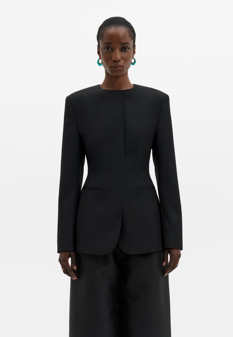 Femme portant un blazer noir structuré avec des épaules rembourrées et un pantalon large assorti, debout devant un fond blanc uni.