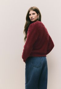 Burgundy, bolyhos pulóver hosszú ujjakkal, magas derekú kék farmernadrággal párosítva. Puha textúra és laza illeszkedés, amelyet a látható varratok hangsúlyoznak.