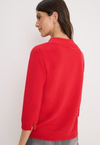 Rode geribbelde top met een hoge kraag, driekwart mouwen en zijsplitten. De textuur is glad met een patroonafwerking. Achteraanzicht getoond.