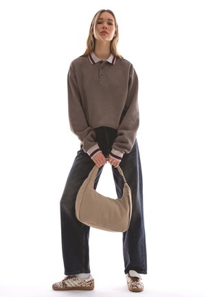 Tânără stând în picioare, purtând un hanorac maro cu guler, blugi închiși la culoare, adidași cu model, ținând cu ambele mâini în față o geantă bej.