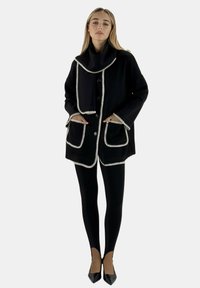 Cappotto nero con rifiniture bianche e ampie tasche, caratterizzato da un colletto ampio e chiusura a bottoni. Indossato sopra leggings neri e scarpe a punta.