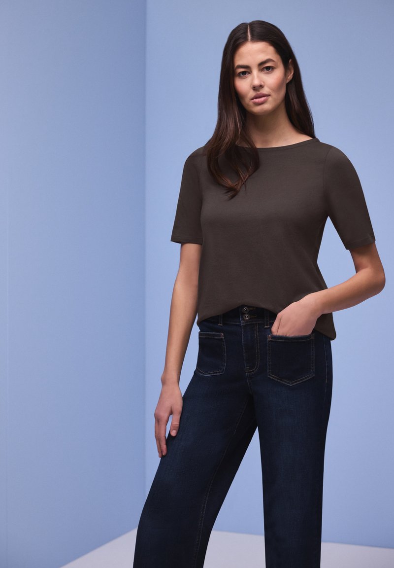 Femme aux longs cheveux bruns se tenant contre un mur bleu clair, portant un haut marron foncé à manches courtes et un jean bleu foncé, une main dans la poche.