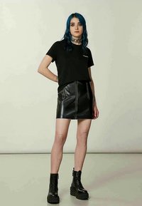 Noor noor sinise juuksevärviga, seljas must t-särk, must nahkne mini-seelik, jämedad mustad saapad ja ahelkett, seistes ühtlase tausta ees.