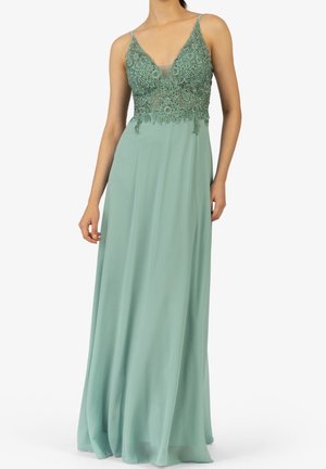 Robe longue sans manches vert pâle avec corsage en dentelle brodée et jupe fluide en chiffon, portée par une personne debout avec les mains détendues.
