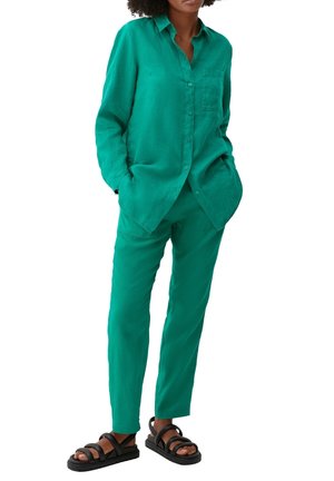 Groen linnen overhemd met knopen, lange mouwen en borstzak, gecombineerd met bijpassende taps toelopende broek. Draagt zwarte sandalen met bandjes.