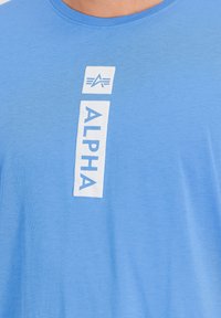 Svetlo modra majica s kratkimi rokavi iz mehkega materiala, ki ima na sredini prsnega dela bel pravokotni logotip z napisom "ALPHA" in grafičnimi poudarki.