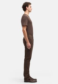 Mężczyzna stojący bokiem, ubrany w brązowy t-shirt, brązowe spodnie i brązowe mokasyny na jednolitym jasnym tle.