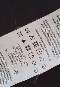 Hoito-ohjeet, joissa on hoitosymboleja, mustaa tekstiä valkoisella kankaalla. Sisältää ohjeet pesemiselle, kuivaamiselle, silitykselle ja materiaaleille.