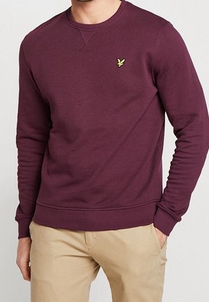Mann trägt einen einfarbigen bordeauxroten Pullover mit Rundhalsausschnitt und kleinem gelbem Vogel-Logo auf der linken Brust sowie beige Hosen, Hand in der Tasche.