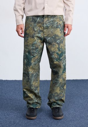 Mand iført grønne og brune camouflagebukser, beige skjorte og sorte sko, stående på blåt tæppe.