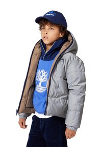 Giacca a vento grigia chiara con cappuccio, fodera marrone, indossata sopra una felpa blu con un logo di un albero bianco, abbinata a pantaloni di velluto a coste scuri.