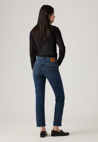 Calças de ganga azul de cintura alta com corte de perna reta, ligeiramente desfiadas na bainha, e um emblema de couro castanho na faixa da cintura traseira.