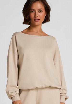 Vrouw met een losse beige lange mouw top met een brede halslijn en bijpassende broek, staand tegen een effen lichte achtergrond.