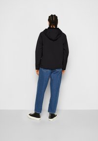 Giacca nera imbottita con cappuccio, dettagli di cucitura visibili, indossata su jeans in denim blu e sneakers nere con suole bianche.