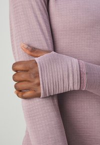Mørkhudet hånd med tommelfinger gennem tommelhul på lys lilla langærmet top, der holder den modsatte arm mod en ensfarvet baggrund.