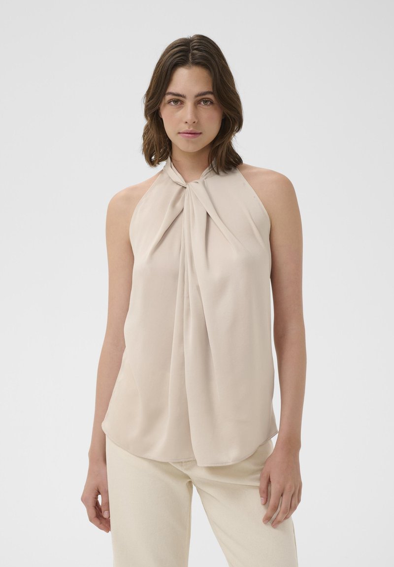 Mlajša ženska z rjavimi ramenskimi lasmi, oblečena v brezrokavnik bež barve in svetle hlače, stoji pred enobarvnim ozadjem.