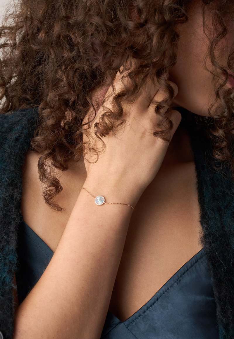 Zachte rosegouden armband met een cirkelvormige stenen centraal, omringd door kleinere heldere stenen, gedragen om een pols tegen een donkere achtergrond.